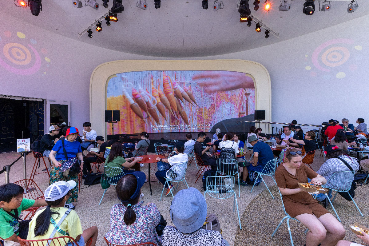 A group of people gather to rest in front of a large screen showing weaving in progress